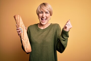 Wall Mural - Young blonde woman with short hair holding fresh bread baguette over yellow background screaming proud and celebrating victory and success very excited, cheering emotion