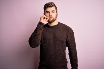 Young blond man with beard and blue eyes wearing casual sweater over pink background mouth and lips shut as zip with fingers. Secret and silent, taboo talking