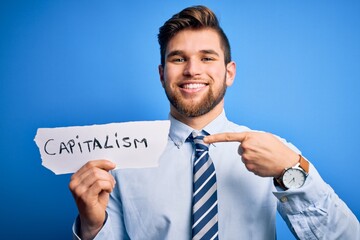 Young blond businessman with beard and blue eyes holding paper with capitalism message very happy pointing with hand and finger