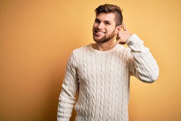 Wall Mural - Young blond man with beard and blue eyes wearing white sweater over yellow background smiling doing phone gesture with hand and fingers like talking on the telephone. Communicating concepts.