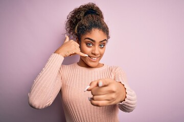 Poster - Young beautiful african american girl wearing casual sweater standing over pink background smiling doing talking on the telephone gesture and pointing to you. Call me.