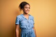 © Krakenimages.com - Young beautiful african american girl wearing denim dress standing over yellow background looking away to side with smile on face, natural expression. Laughing confident.
