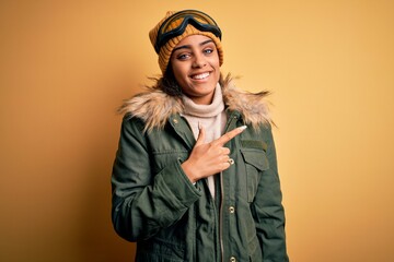 Wall Mural - Young african american afro skier girl wearing snow sportswear and ski goggles cheerful with a smile on face pointing with hand and finger up to the side with happy and natural expression