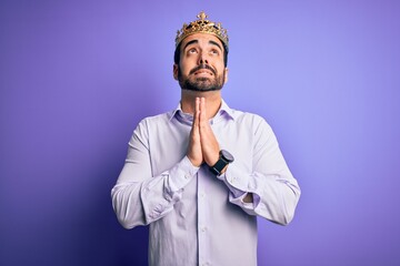 Sticker - Young handsome man with beard wearing golden crown of king over purple background begging and praying with hands together with hope expression on face very emotional and worried. Begging.