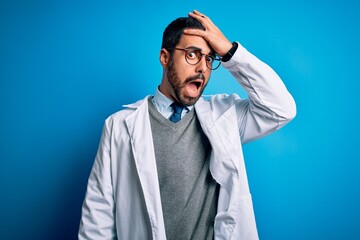 Sticker - Young handsome doctor man with beard wearing coat and glasses over blue background surprised with hand on head for mistake, remember error. Forgot, bad memory concept.