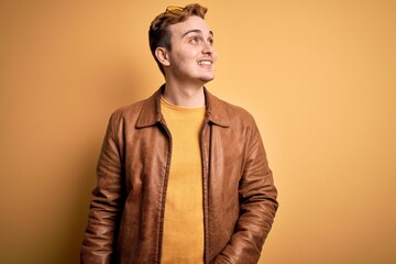 Wall Mural - Young handsome redhead man wearing casual leather jacket over isolated yellow background looking away to side with smile on face, natural expression. Laughing confident.