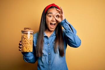 Canvas Print - Young beautiful asian woman holding jar of Italian dry pasta macaroni over yellow background with happy face smiling doing ok sign with hand on eye looking through fingers