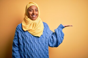 Wall Mural - Young African American afro woman wearing muslim hijab over isolated yellow background smiling cheerful presenting and pointing with palm of hand looking at the camera.