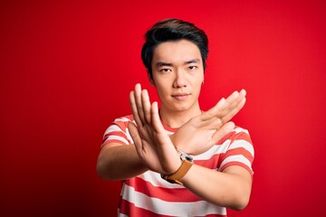 Wall Mural - Young handsome chinese man wearing casual striped t-shirt standing over red background Rejection expression crossing arms doing negative sign, angry face