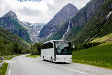 Blue Ice Tongue Of Jostedal Glacier Melts From The Giant Rocky Mountains Into The Green Valley With Waterfalls. Big White Tourist Bus Rides On The Road In Norway In Summer Cloudy Day