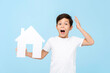 Cute little boy holding home cutout model being shocked and gasping on light blue isolated background