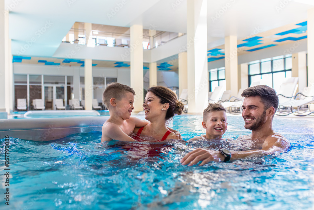 Smiling family of four having fun and relaxing in indoor swimming pool at hotel resort. - obrazy, fototapety, plakaty 