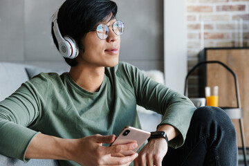 Poster - Image of smiling asian man using cellphone and wireless headphones