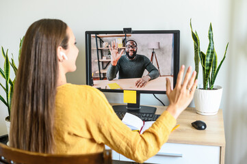 Remote work. A young woman has video meeting with an african-american coworker, employee. She waving hello into webcam. Back view