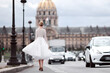 A beautiful young woman walks in a luxurious short white lace dress, high-heeled shoes, next to the road, in Paris, next to the Les Invalides