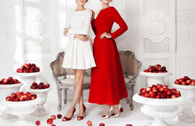 Two Beautiful Young Girls Models, Posing In Luxurious White And Red  Dresses In The Studio, Amid Scenery With Openwork Moldings, A Carved Sofa, White Vases With Red Apples