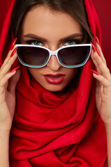 Wall Mural - Portrait of classy brunette young woman with luxurious make-up in white sunglasses and red headscarf on red background. woman look at the camera