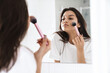 Photo of pleased woman using powder brush while looking at mirror