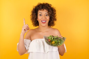 Wall Mural - Charming carefree woman with positive expression, points up with both index fingers, dressed in casual clothing, has broad interested smile, isolated over yellow background. Look there, please.