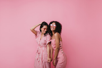 Wall Mural - Excited european girl playing with her hair during photoshoot with friend. Studio portrait of blithesome ladies in pink clothes