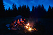 © anatoliy_gleb - Evening camping near fire, forest on background. Group of four friends hikers sitting together near campfire, enjoying fresh air near tent with tourists equipment nearby, dark relaxing atmosphere.