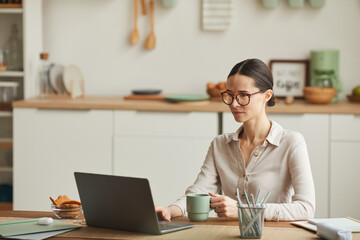 Portrait of elegant young woman drinking coffee while using laptop at cozy home office workplace, copy space