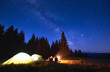 © anatoliy_gleb - Night camping with bonfire. People bask in campfire near illuminated tent city, enjoying valley of mountains in pine forest. Dark blue night sky is strewn with bright stars and Milky Way