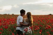 Young Couple Enjoying The Time Together In The Red Meadow. Young Happy Couple Hugging On The Poppy Field. Couple In Love. Beginning Of A Love Story. Red Flowers Background. Copy Space.