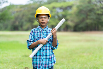 African American boy holding white blueprints and wearing yellow safety helmet while standing outdoor and looking to camera. Education or Field trip concept