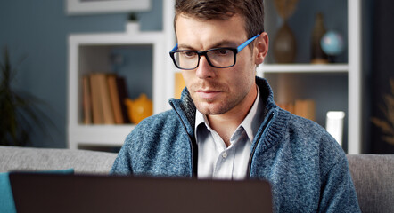 Focused man in eyewear and casual clothes using laptop looking at screen sitting on sofa in apartment