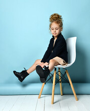 Curly Kid In Beige Dress, Black Jacket And Boots. She Is Posing Sitting On White Chair Against Blue Studio Background