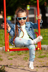 Wall Mural - fashionable child girl in a jeans suit and sunglasses swinging on a swing