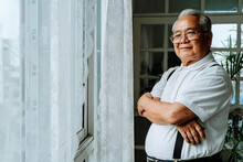 Positive Plump Senior Asian Male Smiling For Camera And Crossing Arms While Resting Near Window In Cozy Room At Home