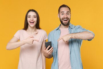 Excited young couple two friends guy girl in pastel blue casual clothes posing isolated on yellow wall background. People lifestyle concept. Mock up copy space. Pointing index fingers on mobile phone.