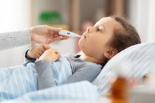 Family, Health And People Concept - Mother With Sick Little Daughter Lying In Bed With Oral Thermometer And Measuring Temperature At Home