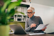© bnenin - Senior woman doing finance at home, holding paper document and looking at laptop.