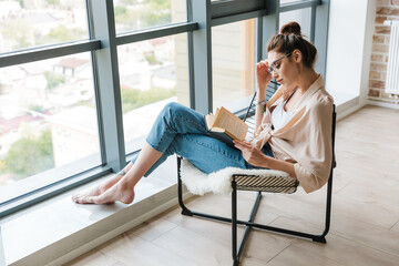 Wall Mural - Image of focused woman reading book while sitting in armchair