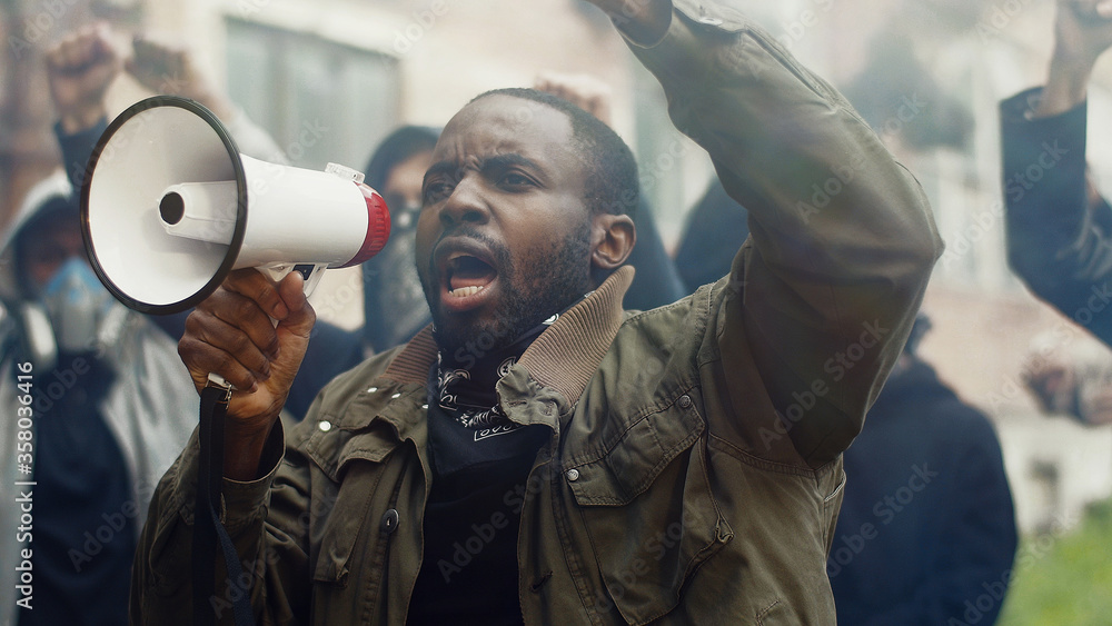 African-American young handsome man screaming in megaphone at protest for human rights outdoors in smoke. Group of people protesting at street. Strike against violence. - obrazy, fototapety, plakaty 