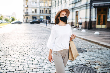 Stylish young woman in protective mask on the face walking in the downtown. Girl in sunglasses and hat in the city during pandemic, epidemic