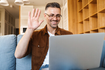 Sticker - Image of cheerful adult man waving hand and working with laptop while