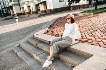 Young woman in trendy wear sits and rests on the step in the downtown. Summer walk in the city