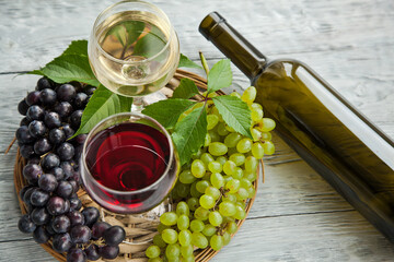 A cluster of dark and green grapes and two wine glasses with red and white wine, bottle on a round straw tray and grey wooden table. Wine composition flat lay.