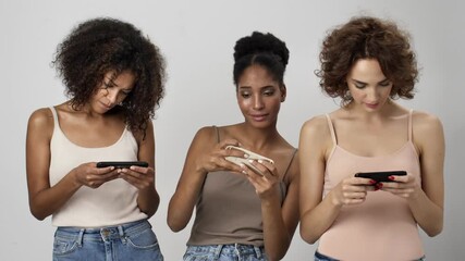 Wall Mural - three attractive multi-ethnic women are playing games on their smartphones standing isolated over wh