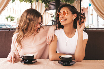 Wall Mural - Two young beautiful smiling hipster girls in trendy summer sundress.Carefree women chatting in veranda cafe and drinking coffee.Positive models having fun and communicating