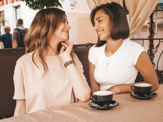 Two young beautiful smiling hipster girls in trendy summer sundress.Carefree women chatting in veranda cafe and drinking coffee.Positive models having fun and communicating