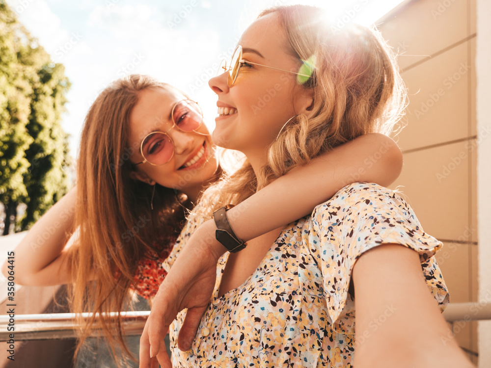 Two young beautiful smiling hipster girls in trendy summer sundress.Sexy carefree women posing on the street background in sunglasses. They taking selfie self portrait photos on smartphone at sunset - obrazy, fototapety, plakaty 