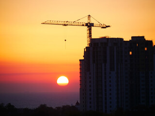 Wall Mural - Construction site background. Silhouette of building under construction against the sea at sunrise. Self-erection crane.