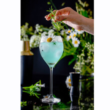 Blue Gin In A Tall Glass Covered By Water Droplets With Summer Daisy Flowers As A Garnish And On A Background.