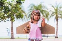 Happy Asian Funny Child Or Kid Little Girl Smile Wear Pilot Hat And Goggles Play Toy Cardboard Airplane Wing Flying Against Summer Sky Cloud On Trees Garden Background, Startup Freedom Concept