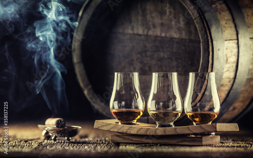 Glencairn whiskey tasting cups on a wooden serving, with a whisky barrel in the dark background and a smoking cigar next to it